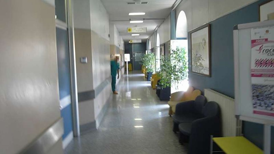 Corridoio interno di un ospedale con pavimento lucido, pareti bicolori e insegne a soffitto, tra cui “SALA CHIRURGICA”. Lungo il lato destro ci sono sedute d’attesa, piante in vaso e grandi finestre ad arco con luce naturale.