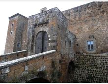 Ingresso di un castello in pietra con portale ad arco e grande porta in legno, raggiunto da un ponte in muratura. Le mura esterne massicce mostrano una torre laterale e una piccola finestra incorniciata in pietra.