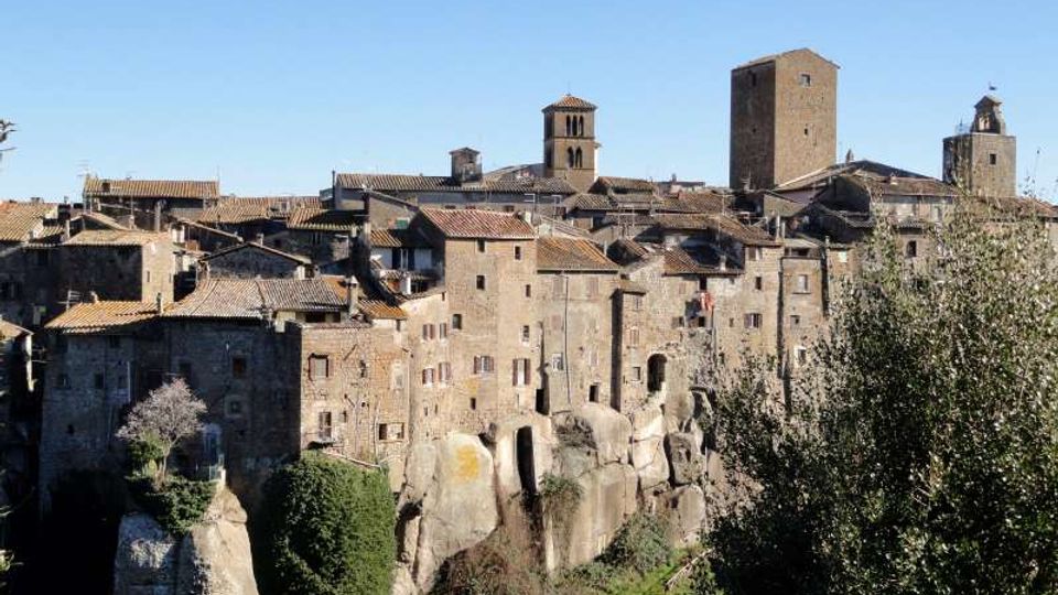 Borgo medievale in pietra arroccato su uno sperone di roccia, con case addossate e tetti in tegole. Sullo sfondo si distinguono una torre quadrata e campanili, con vegetazione in primo piano e cielo sereno.
