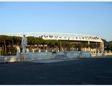 Veduta esterna di uno stadio con struttura ad anello e tralicci bianchi, sullo sfondo di un filare di pini. In primo piano si vede un’ampia piazza asfaltata con gradinate in pietra e una statua su piedistallo.