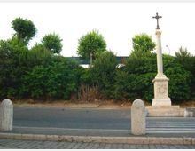 Piazzale asfaltato con cordolo in pietra e alcuni paracarri, davanti a una fitta fascia di vegetazione e alberi. A destra si trova un monumento in pietra con basamento e colonna sormontata da una croce.