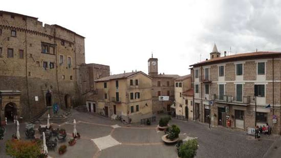 Piazza lastricata in un centro storico, circondata da edifici in pietra con persiane e portici, e dominata a sinistra da un grande edificio fortificato medievale con scalinata. Sullo sfondo si vede una torre con orologio e al centro della piazza sono presenti fioriere e arredi urbani.