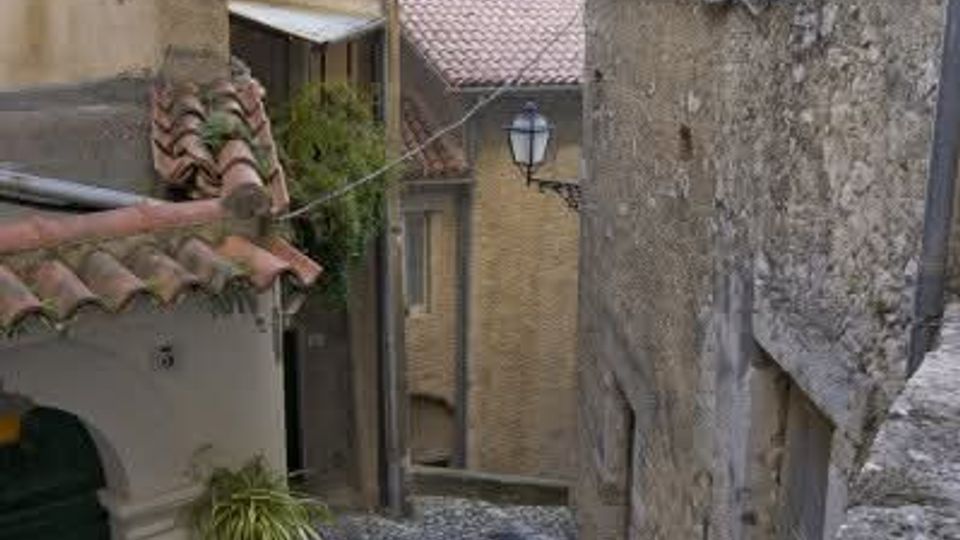 Stretta viuzza in discesa lastricata in pietra, tra edifici in muratura e pietra con tetti in coppi. Un lampione a parete e cavi sospesi attraversano il vicolo, con piccoli ingressi e balconcini affacciati sulla strada.