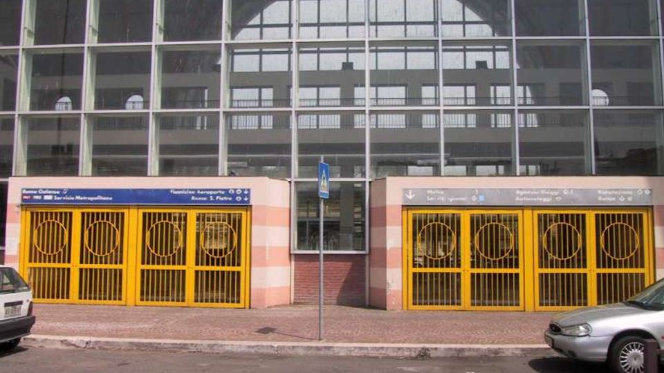 Ingresso di una stazione della metropolitana sotto una grande facciata vetrata a griglia, con due varchi affiancati chiusi da cancelli gialli e pannelli di segnaletica sopra le aperture. Davanti si vedono marciapiede e strada con auto parcheggiate.