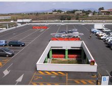 Grande parcheggio asfaltato con strisce bianche e numerosi posti auto, con alcune auto parcheggiate e aree con carrelli della spesa sotto pensiline. In primo piano si vede una struttura rettangolare incassata con pareti bianche e pilastri verdi, mentre sullo sfondo ci sono campi aperti e colline lontane.