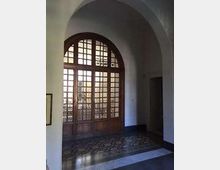 Interno di un edificio con ampia porta finestra ad arco in legno e vetri a riquadri, affacciata su un cortile esterno. Pavimento a mosaico con motivi geometrici e soffitto alto con volta ad arco.