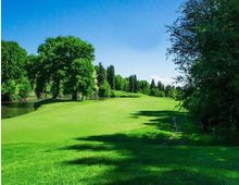 Ampio prato verde ondulato in un parco, costeggiato da un piccolo specchio d’acqua sulla sinistra e circondato da alberi ad alto fusto. In primo piano si vede un sentiero stretto tra l’erba, con cielo sereno sullo sfondo.