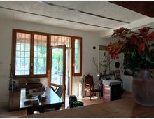 Interno di una sala da pranzo rustica con soffitto a travi, grande tavolo in legno e ampie finestre e porta finestra con griglie che si aprono su un patio esterno. Arredi e oggetti decorativi sono distribuiti lungo le pareti, con un grande vaso di fiori in primo piano.