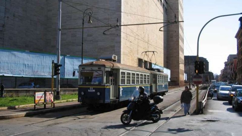 Strada urbana con binari del tram al centro e cavi aerei, con un tram blu in transito accanto a un grande edificio moderno in cemento. Sul lato destro si vedono auto in corsia e lampioni e semafori lungo il marciapiede.