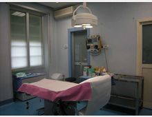 Sala visite medica con lettino coperto da teli bianchi e rosa, lampada chirurgica a soffitto e carrello con strumenti e materiali sanitari. Pareti azzurre con finestra, condizionatore e apparecchiature di monitoraggio fissate al muro.