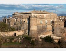 Grande edificio storico in pietra, simile a un castello o palazzo fortificato, con torre cilindrica, mura alte e finestre rettangolari. In primo piano si vede una rampa murata con arco d’ingresso, vegetazione e rovine, con colline sullo sfondo sotto un cielo nuvoloso.