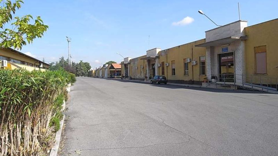 Strada ampia e asfaltata in un’area industriale, con una fila di edifici bassi gialli sulla destra dotati di ingressi, finestre e condizionatori esterni. A sinistra si vede una siepe lungo il marciapiede, con lampioni e alberi sullo sfondo sotto un cielo sereno.
