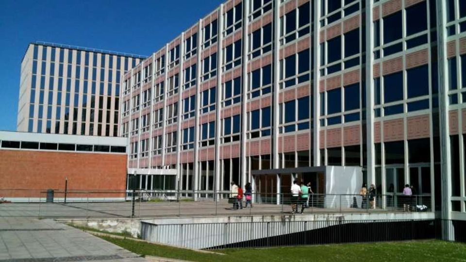Complesso di edifici moderni per uffici o università con facciate a griglia di grandi finestre e pannelli in laterizio, affacciato su un’ampia area pavimentata con prato. In primo piano si vede una rampa o accesso ribassato con ringhiere metalliche sotto un cielo sereno.
