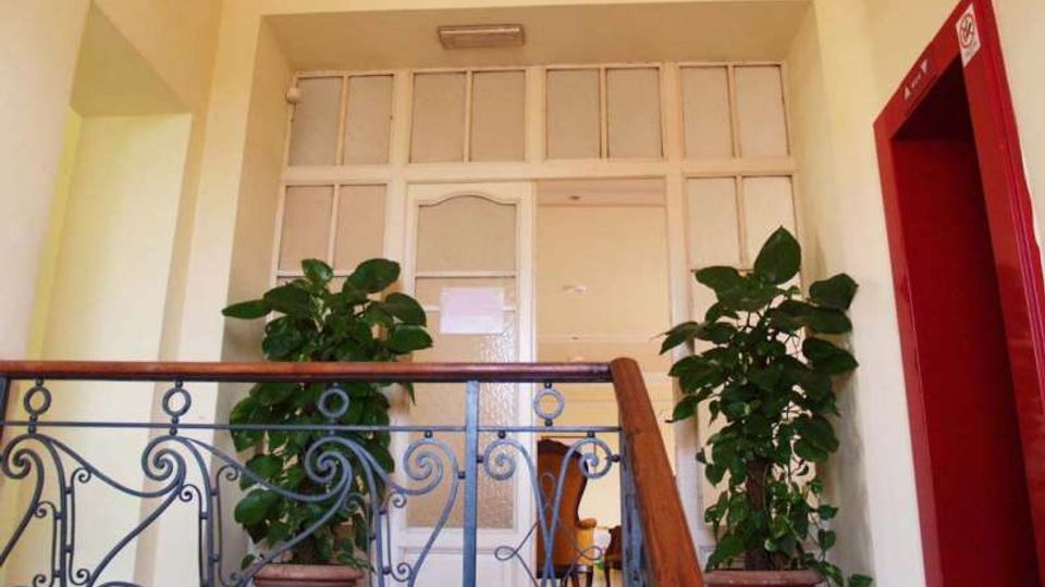 Interno di un edificio con pianerottolo e ringhiera in ferro battuto, pareti chiare e porta vetrata con finestre superiori. Ai lati della porta ci sono due grandi piante in vaso e sulla destra un vano o porta aperta con cornice rossa.