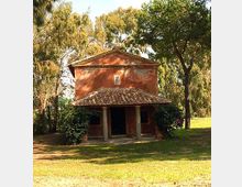 Piccola chiesa rurale in muratura intonacata color rosso, con facciata a timpano e portico d’ingresso con due colonne e tetto in coppi. L’edificio è immerso in un’area verde alberata con prato e alti pini tutt’intorno.