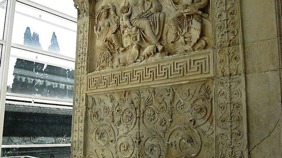 Grande pannello in pietra con bassorilievi figurativi e motivi floreali intrecciati, incorniciato da decorazioni scolpite e fregi a greca. L’opera è esposta in un ambiente museale con parete vetrata e soffitto in vetro che lasciano intravedere l’esterno.