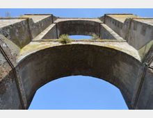 Veduta dal basso di una struttura in pietra tipo ponte o acquedotto con grandi arcate sovrapposte e pareti laterali massicce. Sulla sommità si notano vegetazione e tracce di muschio, con cielo azzurro sullo sfondo.