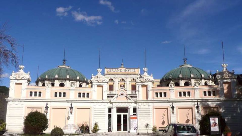 Facciata simmetrica di un grand hotel in stile classico, con due grandi cupole verdi laterali e ingresso centrale decorato con frontone e colonne. Davanti si vede un piazzale con posti auto, aiuole e lampioni, sotto un cielo sereno.