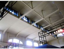 Vista verso l’alto dell’interno di un grande edificio industriale con soffitto alto a capriate e travi in cemento, finestre a nastro lungo la sommità e archi sulle pareti. Sono presenti strutture metalliche a griglia e un’illuminazione con faretti sospesi.