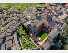 Veduta aerea di un borgo storico con un grande castello in pietra dotato di torri merlate, mura e un ampio cortile interno con prato e fontana. Intorno si vedono case con tetti in coppi e una strada che costeggia i giardini geometrici all’interno delle fortificazioni.