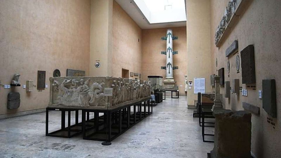 Sala museale con pareti alte color beige e lucernario rettangolare sul soffitto, pavimento in pietra e reperti lapidei esposti lungo le pareti. Al centro ci sono grandi bassorilievi su supporti metallici in fila e, in fondo, una colonna monumentale su piedistallo.