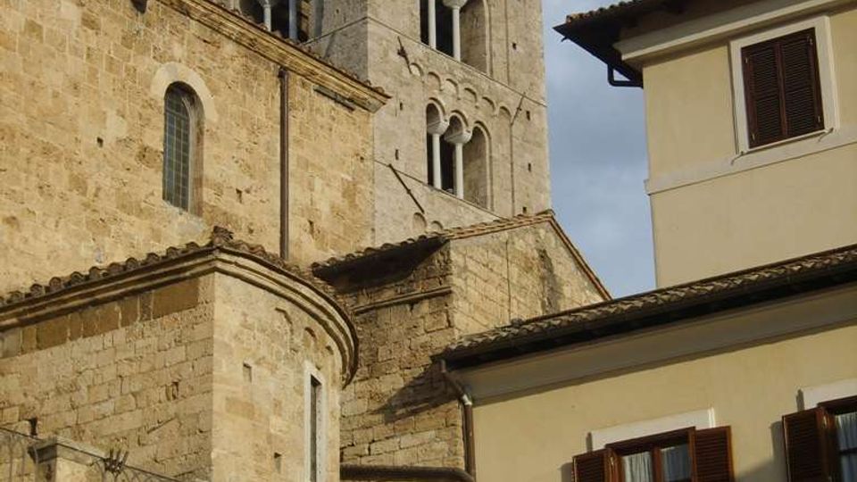 Torre campanaria in pietra di una chiesa in stile romanico, con finestre ad arco e muratura a blocchi, affiancata da un edificio residenziale chiaro con persiane in legno. In basso si vede un’arcata in pietra e, sullo sfondo, il cielo parzialmente nuvoloso.