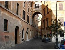 Stretta via urbana tra edifici storici in intonaco chiaro, con finestre a persiane e portoni ad arco al piano terra. In fondo si vede un grande arco che collega i palazzi sopra la strada, con un lampione e alcune auto parcheggiate lungo il bordo.