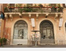 Facciata di un edificio storico con due grandi archi al piano terra chiusi da serrande e grate decorative, e una balconata superiore con ringhiera in ferro e molte piante in vaso. Al centro si vede una piccola fontanella su un marciapiede in pietra.