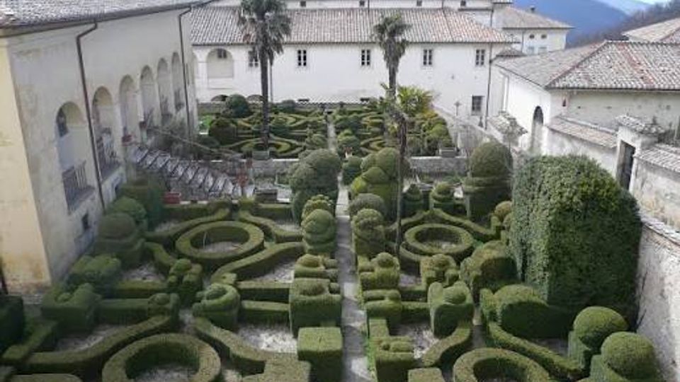 Veduta dall’alto di un chiostro con giardino formale all’italiana, caratterizzato da siepi potate in forme geometriche e vialetti centrali. Il cortile è circondato da edifici in muratura chiara con tetti in coppi e un campanile sullo sfondo, con montagne in lontananza.