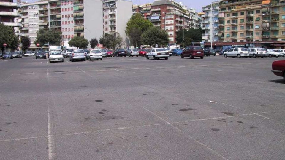 Ampio parcheggio asfaltato con segnaletica a terra e numerose auto, circondato da palazzi residenziali multipiano con balconi. Sullo sfondo si vedono alberi lungo il margine del piazzale sotto un cielo sereno.