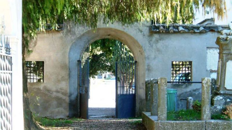 Ingresso con arco in muratura in un muro intonacato, con cancello metallico aperto che conduce a un’area esterna alberata. Ai lati dell’arco si vedono piccole finestre con grata e un vialetto di ghiaia sotto rami pendenti.