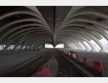 Interno di un grande viadotto o sottopasso in cemento con archi ripetuti e soffitto a volte, visto in prospettiva verso un’apertura luminosa. Sul pavimento si vedono fasci di cavi o barre stesi longitudinalmente e barriere stradali rosse e bianche ai lati.