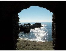 Veduta del mare e di una piccola spiaggia sabbiosa con scogli affioranti, incorniciata dall’apertura ad arco di una struttura in mattoni. L’acqua è illuminata dal riflesso del sole, con l’orizzonte visibile in lontananza.