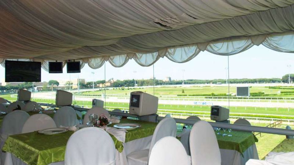 Sala hospitality con soffitto drappeggiato in tessuto e tavoli apparecchiati, con sedie rivestite di bianco e tovaglie verdi. Grandi vetrate affacciano su un ippodromo con pista e prato, con schermi appesi e piccoli monitor lungo il parapetto.