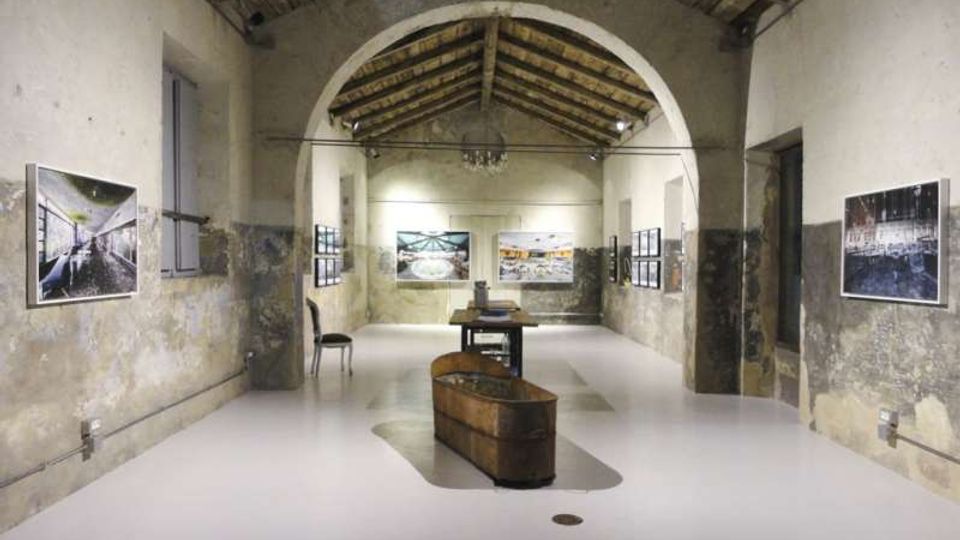 Interno di un edificio in stile industriale riconvertito a spazio espositivo, con soffitto a capriate in legno e grande arco centrale. Pareti intonacate e parzialmente scrostate con fotografie appese, pavimento chiaro e tavoli con oggetti al centro della sala.