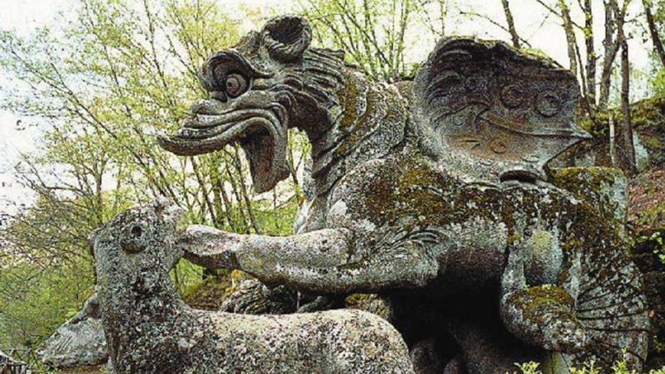 Grande scultura in pietra ricoperta di muschio raffigurante una creatura alata con becco e una figura animale allungata, situata in un’area boschiva con alberi e vegetazione sullo sfondo. La scena appare come un gruppo scultoreo all’aperto, integrato tra rocce e cespugli.
