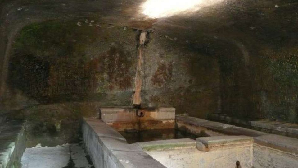 Interno di una grotta o struttura in pietra con vasche rettangolari in muratura piene d’acqua e una piccola fonte centrale che sgorga dal muro. Una luce dall’alto illumina parzialmente le pareti scure e umide.