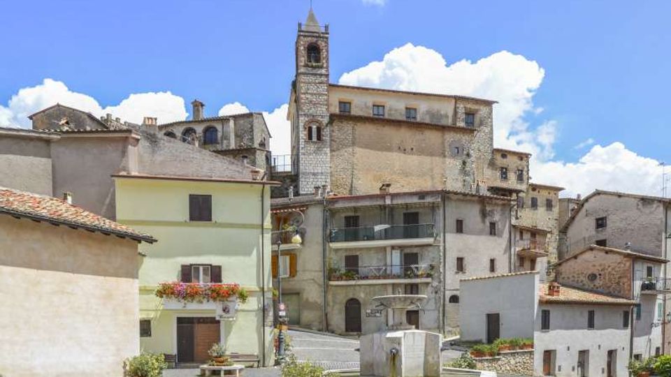 Piazza di un borgo storico con pavimentazione in pietra a motivi curvi e una fontana centrale. Sullo sfondo si vedono case in muratura e un campanile in pietra che svetta tra edifici con tetti in coppi, sotto un cielo azzurro con nuvole.