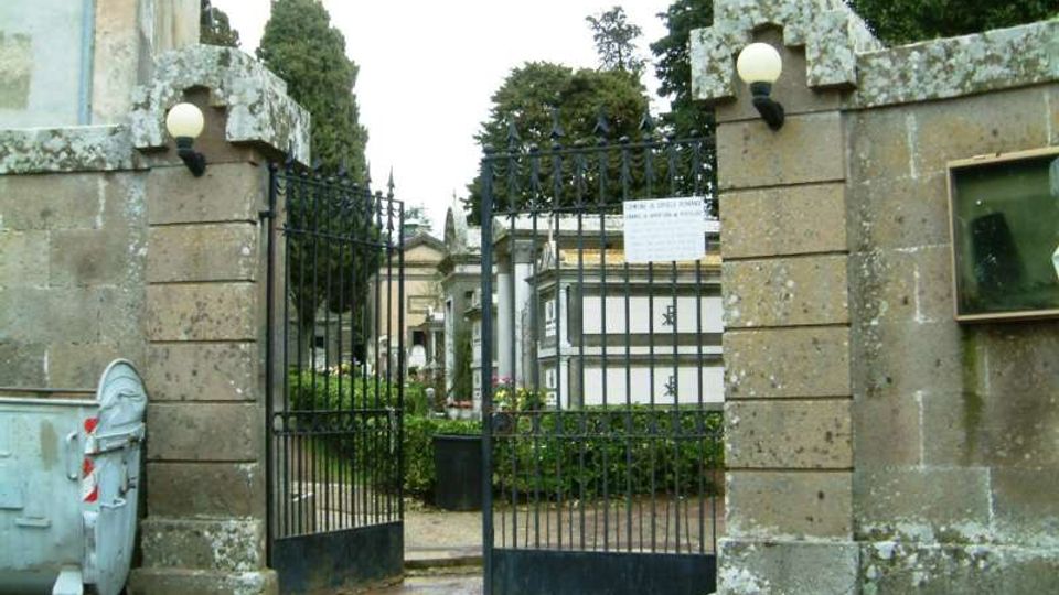 Ingresso di un cimitero con cancello in ferro battuto tra due pilastri in pietra con lampade sferiche. Sullo sfondo si vedono cappelle e tombe tra alberi e siepi, con un percorso interno in ghiaia.