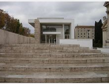 Edificio moderno con facciata vetrata e volume bianco sporgente, raggiungibile tramite un’ampia scalinata in pietra. Ai lati si vedono muri in travertino, alberi e altri edifici in un contesto urbano sotto cielo nuvoloso.