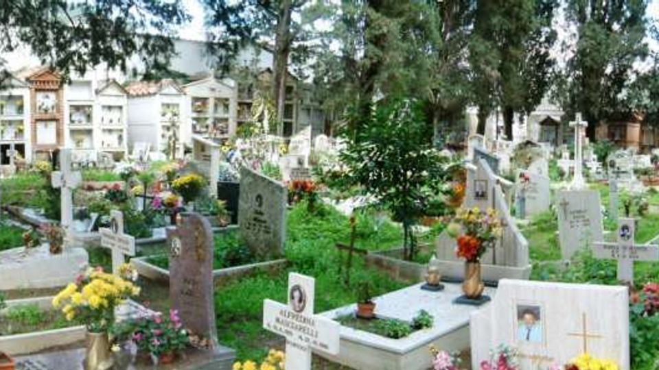 Cimitero all’aperto con numerose lapidi e tombe decorate con fiori, immerse nel verde e ombreggiate da alti alberi. Sullo sfondo si vedono loculi e cappelle in muratura, con un vialetto pavimentato lungo il lato destro.