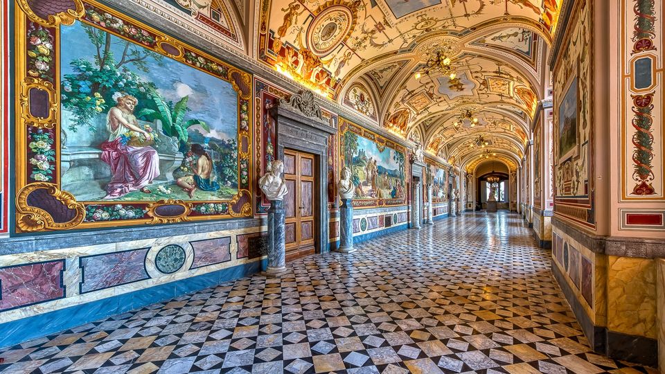 Lungo corridoio interno di un palazzo storico, con pavimento a mosaico geometrico in marmo e pareti rivestite da grandi affreschi incorniciati e decorazioni policrome. Il soffitto a volta è riccamente ornato con dipinti e stucchi, illuminato da lampadari, con porte in pietra e statue lungo il percorso.