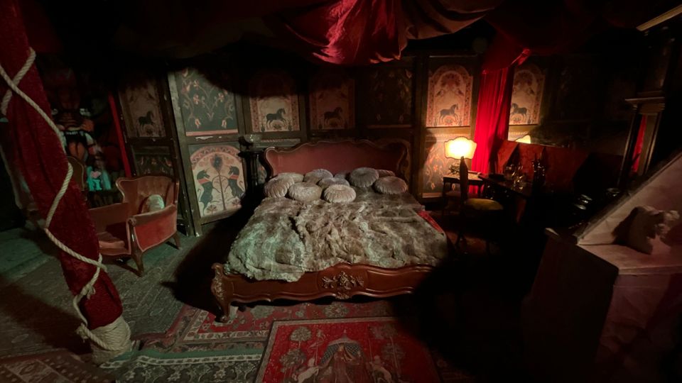 Interno di una camera da letto in stile antico con grande letto in legno e coperta di pelliccia, cuscini decorativi e tappeti ornamentali. Le pareti sono rivestite con pannelli dipinti e tende rosse drappeggiate, con lampade soffuse e mobili vintage ai lati.