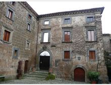Cortile interno di un edificio storico in pietra con facciata a più piani, finestre con scuri in legno e archi in muratura. Si vedono un grande portone ad arco con gradini in pietra, un accesso ad arco più piccolo e pareti parzialmente coperte da rampicanti.