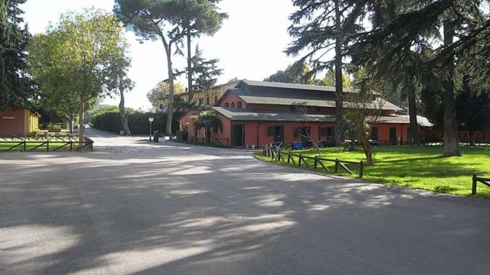 Ampio viale asfaltato in un’area verde con prati, staccionate basse e alti alberi; sullo sfondo si vede un edificio basso con facciata rosa e tetto scuro, affacciato sulla strada.
