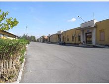 Strada ampia e asfaltata in un’area industriale, con una fila di edifici bassi gialli sulla destra dotati di ingressi, finestre e condizionatori esterni. A sinistra si vede una siepe lungo il marciapiede, con lampioni e alberi sullo sfondo sotto un cielo sereno.