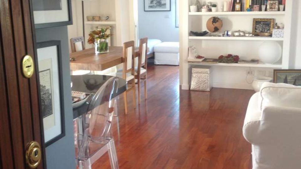 Interno di un soggiorno-pranzo con pavimento in legno, librerie bianche a parete piene di libri e oggetti decorativi. A sinistra c’è un tavolo da pranzo con sedie e uno sgabello trasparente, a destra un divano bianco, con una porta centrale che conduce a una stanza luminosa.