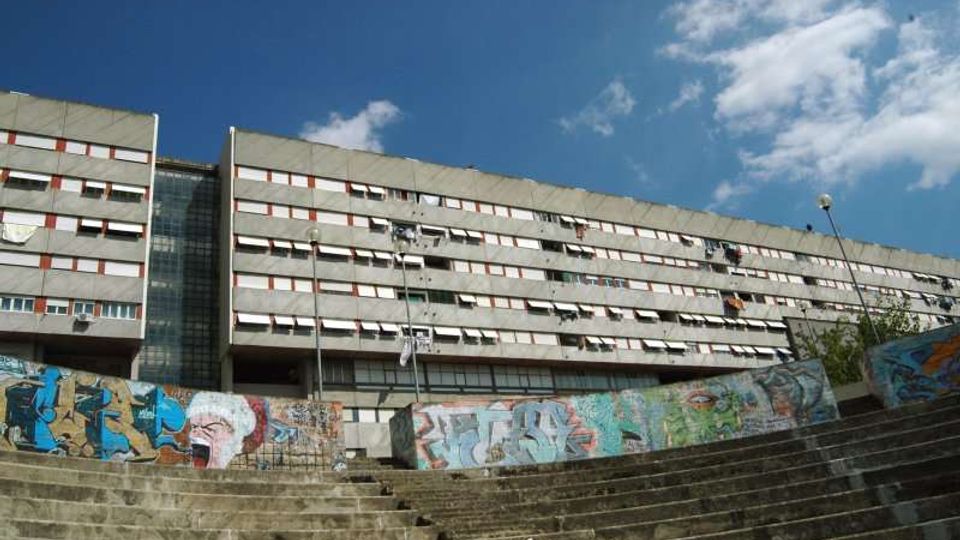 Grande edificio residenziale a più piani con facciata in pannelli grigi e lunghe file di finestre, con una torre vetrata centrale. In primo piano ci sono ampie gradinate in cemento e un muro basso coperto di graffiti, sotto un cielo azzurro con nuvole.