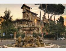 Fontana monumentale in una rotonda stradale, con grande vasca circolare e sculture in pietra alla base. Sullo sfondo si vede un edificio storico in stile liberty con torre e facciata decorata, circondato da alti pini e vegetazione urbana.