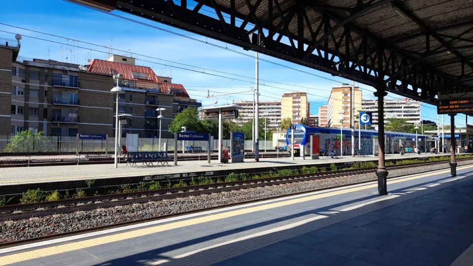 Banchine della stazione ferroviaria Roma Trastevere con binari multipli e pensilina in ferro, illuminate dal sole. Sullo sfondo si vedono un treno regionale fermo, cartelli di stazione, lampioni e palazzi residenziali.